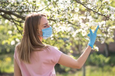 Coronavirus. Yüzü maskeli kadın karantinada. Evde kal. Evde yemek pişirmenin tadını çıkar. Aile kavramı. Genç bir kadın karahindiba üflüyor. Sahilde bir bardak şampanya. Suyla oynayan kadın. Kadın şişeden su içiyor.