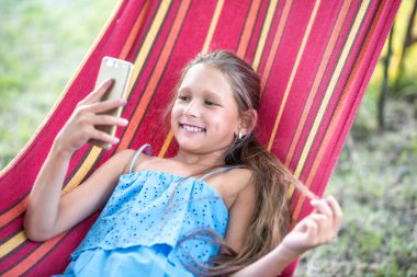 Güzel bir kız hamakta dinleniyor. Kız telefonda selfie çekiyor. Aile memnuniyeti. Bir yaz günü dinlenin. gülümseyen çocuk.