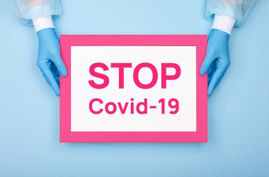 Hastalığın koronavirüse karşı temasının gerçek fotoğrafı. Tıbbi eldivenlerdeki eller mavi arkaplanda COVID-19 yazan posteri tutuyor..