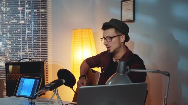 Joven barbudo con sombrero cantando emocionalmente en el micrófono y tocando la guitarra en el estudio en casa — Vídeos de Stock