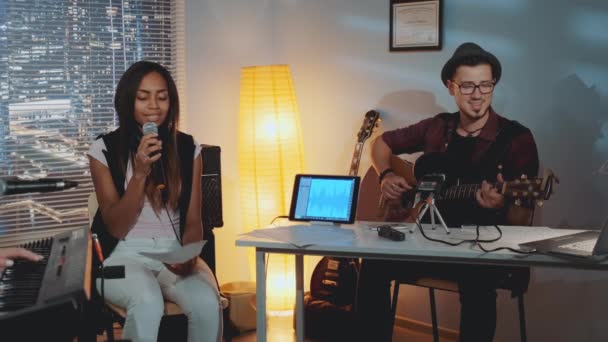 Modern band rehearsing in home studio: young man playing guitar and mixed-race girl singing into microphone — Stock Video