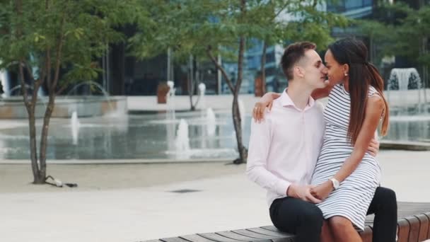 Dos amantes de las carreras mixtas sentados en un banco y besándose — Vídeo de stock