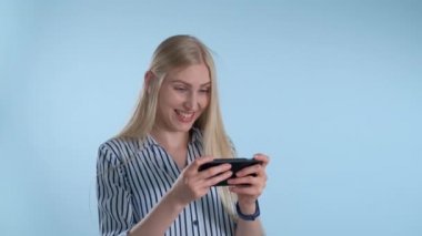 Güzel sarışın kız mavi arka planda akıllı telefondan oyunlar oynuyor.