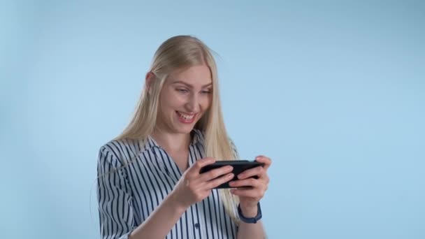 Chica rubia bonita jugando juegos en el teléfono inteligente sobre fondo azul — Vídeo de stock