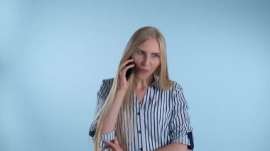 Hoş görünümlü genç bir bayan mavi arka planda akıllı telefondan biriyle konuşuyor..