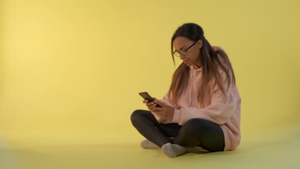 Mulher negra alegre escrevendo mensagem no smartphone sentado no chão . — Vídeo de Stock