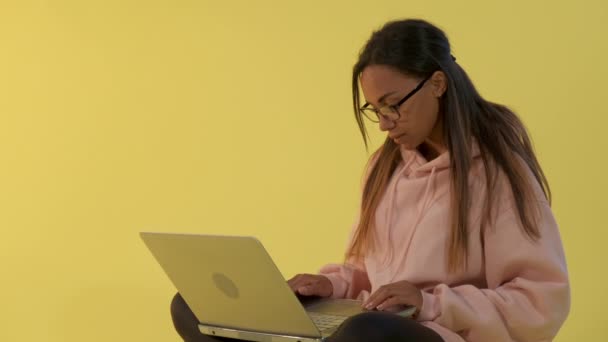 Jovencita de raza mixta trabajando en el ordenador sobre fondo amarillo — Vídeos de Stock