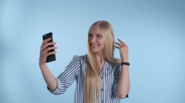 Çekici sarışın bayan mavi arka planda akıllı telefondan selfie çekiyor.