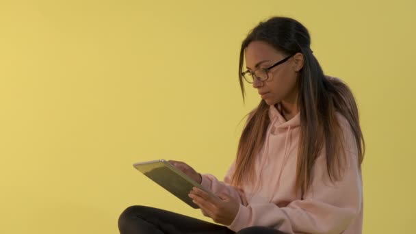 Czarna kobieta w okularach pracująca nad tabletką na żółtym tle. — Wideo stockowe