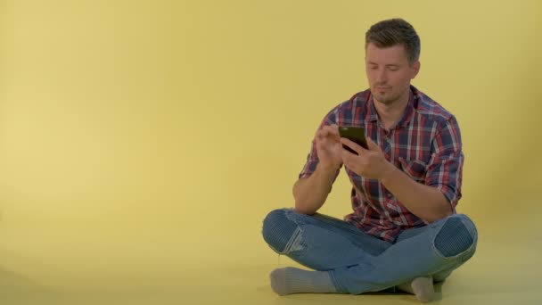 Niño guapo sentado en el suelo y charlando con amigos en el teléfono inteligente — Vídeos de Stock