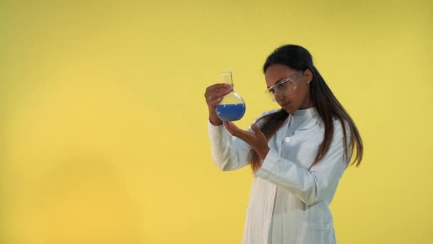 Black female scientist in lab coat looking on flask with experimental liquid on yellow background. — Stock Video