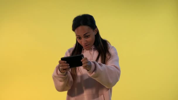 Приємно виглядає чорна дівчина грає в ігри на смартфоні на жовтому фоні . — стокове відео