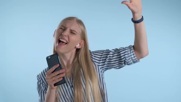 Молода блондинка стає високою, слухаючи музику і співаючи на синьому фоні — стокове відео