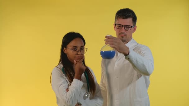 Wieloetniczny mężczyzna i kobieta w fartuchach laboratoryjnych patrzący na kolbę z płynem doświadczalnym na żółtym tle. — Wideo stockowe