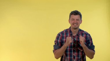 Kareli gömlekli heyecanlı adam sarı arka planda yumruklarını kaldırıyor..