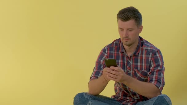 Veselý muž v kostkované košili sedí na podlaze a klábosí na smartphonu. — Stock video
