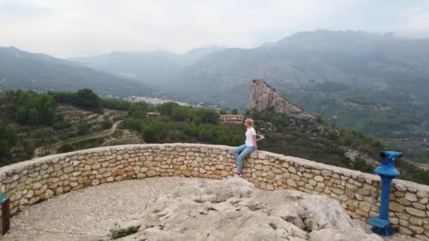 Mulher sentada e olhando para uma bela vista com um lago azul e montanhas a partir da parede de um castelo velho — Vídeo de Stock