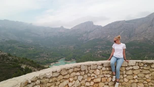 Nő ül, és néz egy gyönyörű kilátás egy kék tó és hegyek a fal egy régi vár — Stock videók
