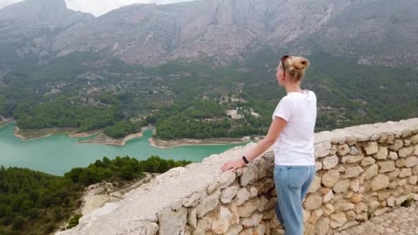 Uma jovem mulher olha para uma bela vista com um lago azul e montanhas a partir da parede de um antigo castelo — Vídeo de Stock