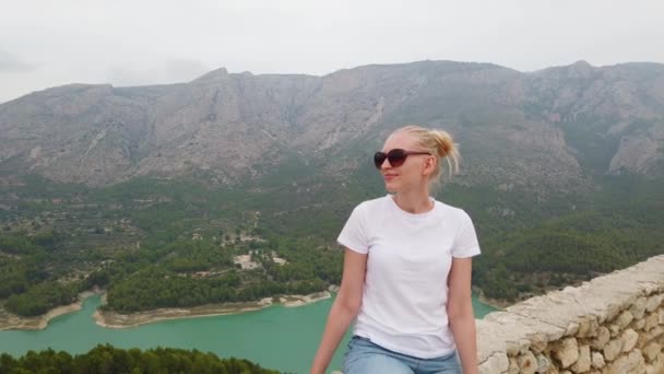 Nő ül, és néz egy gyönyörű kilátás egy kék tó és hegyek a fal egy régi vár — Stock videók