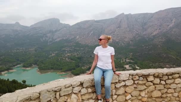 Mulher sentada e olhando para uma bela vista com um lago azul e montanhas a partir da parede de um castelo velho — Vídeo de Stock