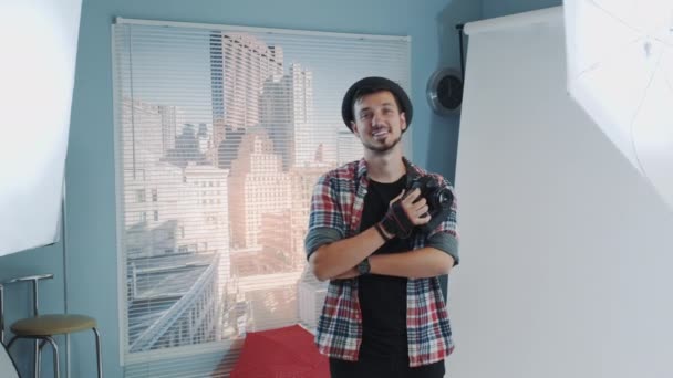 Guapo joven fotógrafo en sombrero sosteniendo la cámara y sonriendo en estudio moderno sesión de fotos — Vídeos de Stock