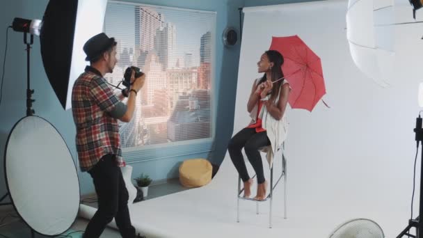 Elegante modelo africano posando con paraguas rojo en la barra de la silla alta para la revista de moda sesión de fotos — Vídeo de stock