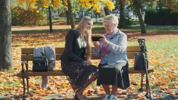 美しいですブロンド女の子とともに年上の女性座っていますベンチと何かを見てスマートフォン — ストック動画