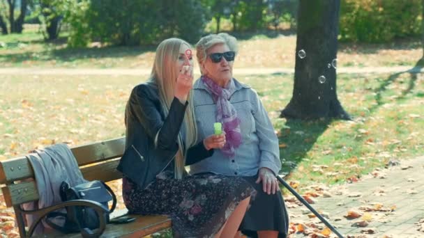Średnie ujęcie Szczęśliwej młodej kobiety dmuchającej bąbelki siedząc razem z babcią w parku — Wideo stockowe
