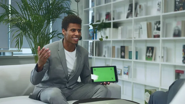 Black formal man making presentation with tablet with green chromakey screen. Young African-American man in suit sitting in office on sofa with anonymous colleague and making presentation with tablet.