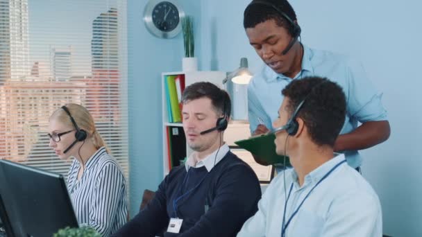Mittlere Aufnahme eines multiethnischen Kundendienstmitarbeiters, der seinen Kollegen einen Witz erzählt, während er die Kunden anruft — Stockvideo