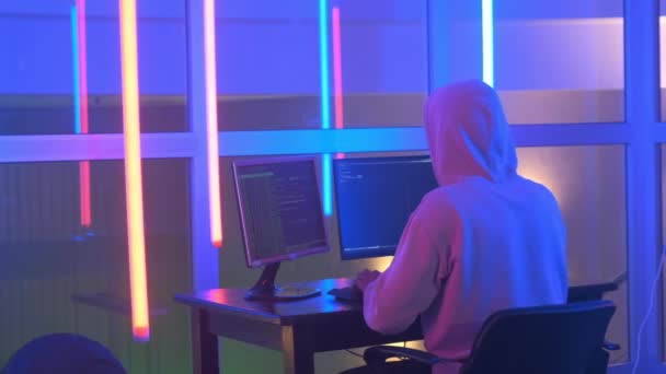 Back view of Hacker in white hoodie penetrating network system in neon room — Stockvideo