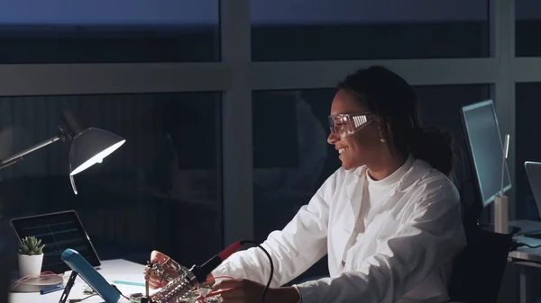 Especialista en electrónica afroamericana en gafas protectoras y bata blanca trabajando con probador de multímetros y placa base en laboratorio — Foto de Stock