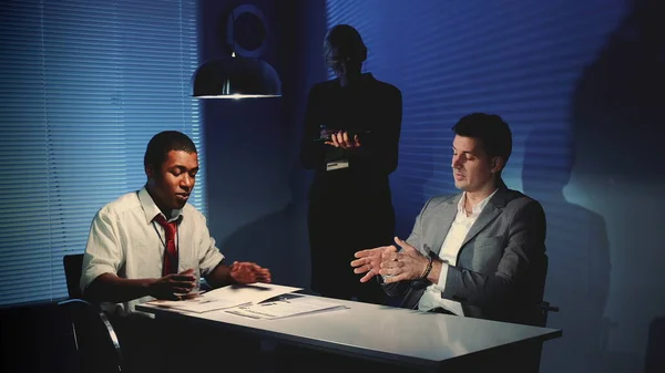 Detective africano interrogando a sospechoso caucásico en sala de interrogatorios . — Foto de Stock