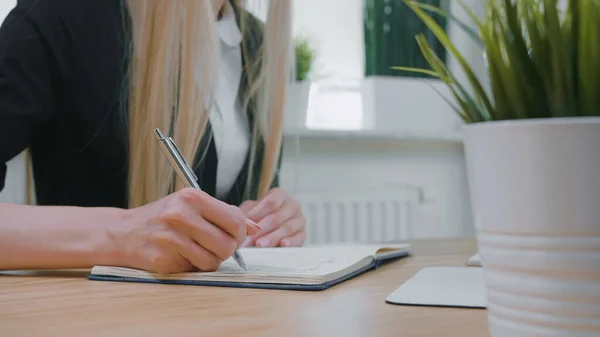 Scrittura femminile con penna in blocco note. Crop vista della donna con lunghi capelli biondi in camicia casual luminoso seduto alla scrivania di legno e annotare le informazioni con penna in metallo lucido nella pianificazione quotidiana di giorno . — Foto Stock