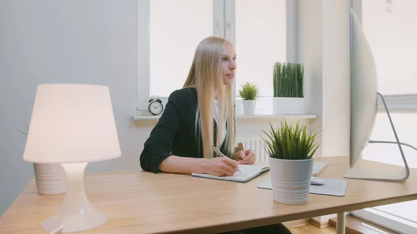 Hermosa mujer de negocios concentrada en el trabajo. Atractiva joven rubia en chaqueta elegante sentada con bolígrafo y portátil en el escritorio con computadora y mirando con entusiasmo a la pantalla del monitor . — Foto de Stock