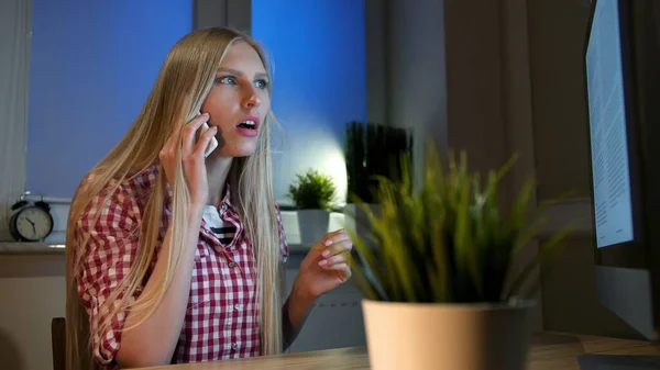 Donna stupita a computer che parla su smartphone. Attraente donna bionda seduta alla scrivania di legno e guardando a bocca aperta il monitor del computer che parla sul telefono cellulare in camera oscura di notte — Foto Stock