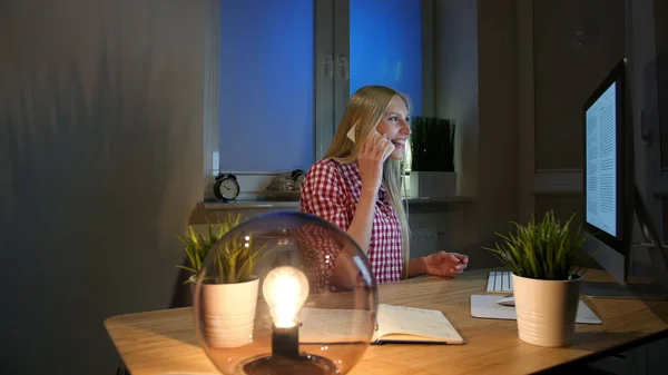 Glückliches Weibchen am Computer im Smartphone-Gespräch. Fröhlich staunende blonde Frau im karierten Hemd sitzt nachts am Computer am Holztisch und telefoniert. — Stockfoto