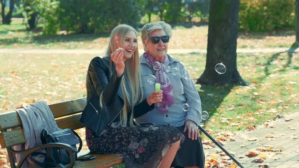 Happy young woman blowing bubbles sitting together with her grandmother in park — 스톡 사진