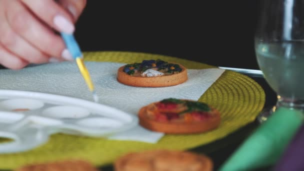 Cookies διακόσμηση τέχνης: γυναίκα ζωγραφική μπισκότα με πινέλο και τα χρώματα των τροφίμων στην παλέτα. 4K — Αρχείο Βίντεο