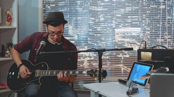 Şapkalı ve modaya uygun kıyafetleri olan gitarist gitar çalıyor ve kayıt stüdyosunda şarkı söylüyor.