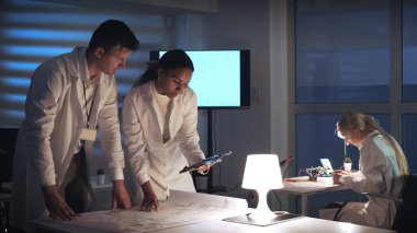 Laboratuvar önlüklü bir adam ve kadın elektronik laboratuarında çalışıyor. Ana kart ve kontrol elektroniği şeması var.