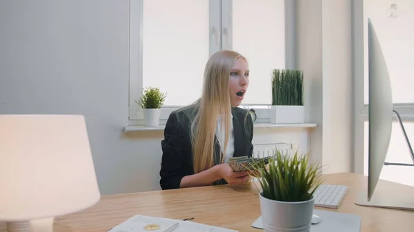 Mujer celebrando el éxito en el cargo. Elegante mujer rubia sentada en el lugar de trabajo sosteniendo en las manos un paquete de dinero en efectivo mirando asombrado al monitor, y tirando dinero en el aire y haciendo alegremente sí gesto . — Foto de Stock