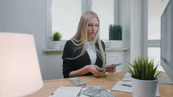 Mujer contando dinero en el lugar de trabajo. Hermosa joven rubia en traje de negocios sentada en la oficina en el escritorio de madera clara con la computadora concentrándose en contar una gran pila de dinero en efectivo en las manos — Foto de Stock