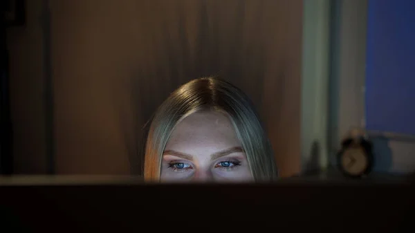 Kobieta podgląda komputer w nocy. Widok zboża pięknej kobiety siedzącej w nocy na komputerze i patrząc na kamerę na górze monitora z ciemnym oknem na rozmytym tle — Zdjęcie stockowe