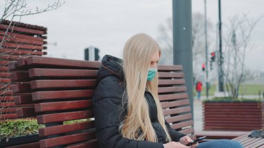 Tıbbi maskeli sarışın kız bankta oturuyor ve akıllı telefon kullanıyor.