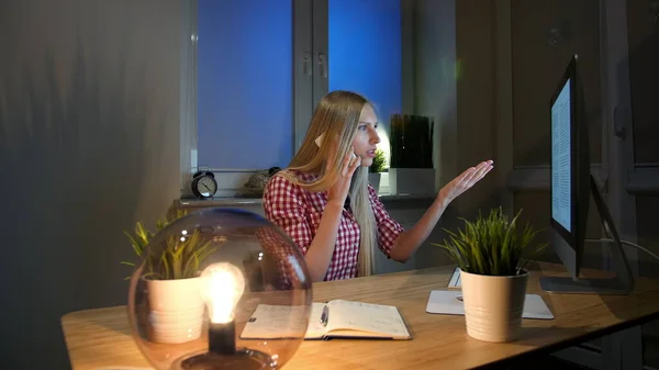 Mujer conmocionada en el ordenador hablando en el teléfono inteligente. Sentado en la habitación oscura en el escritorio de madera con portátil joven rubia hablando en el teléfono móvil y mirando asombrado en el monitor de la computadora. — Foto de Stock