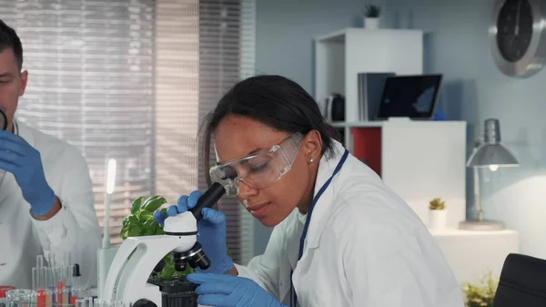Mujer de raza mixta en bata de laboratorio y gafas de seguridad trabajando con microscopio — Foto de Stock