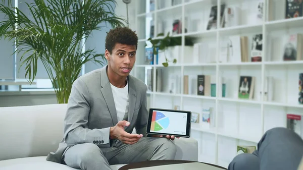 Black formal man making presentation with tablet. Young African-American man in suit sitting in office on sofa with anonymous colleague and making presentation with tablet.