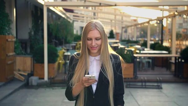 Formal business woman walking on street. Elegant blond young woman in suit and walking on street and browsing smartphone with smile against urban background.